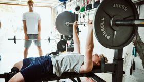 Young man doing bench presses while working out with friends in gym in garage