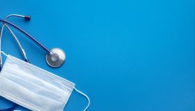 Protective Disposable Medical Face Mask, Stethoscope And Thermometer On A Blue Background.
